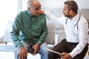 Healthcare worker giving bad news with hand on shoulder of male patient. Doctor consoling sad senior man in waiting room. They are sitting at hospital.