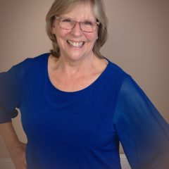 A therapist at Total Life dressed in a vibrant blue blouse, radiating warmth and positivity with her bright smile. She has short, light hair and wears glasses, exuding an approachable and professional demeanor. The neutral background enhances her friendly presence, making her appear welcoming and supportive.