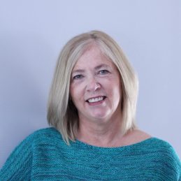 A therapist at Total Life is shown smiling, wearing a teal sweater, and positioned against a soft, neutral background, creating a warm and welcoming impression.