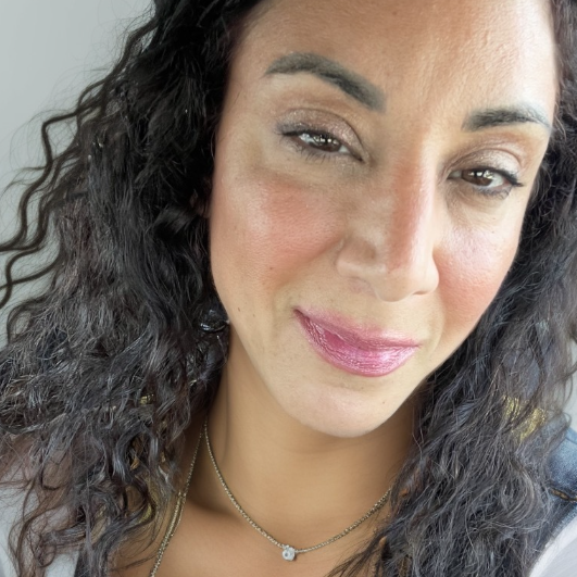 A therapist at Total Life with curly dark hair, a kind smile, and soft makeup featuring a natural look. She is wearing a delicate necklace and is photographed in natural light, creating a warm and inviting appearance.