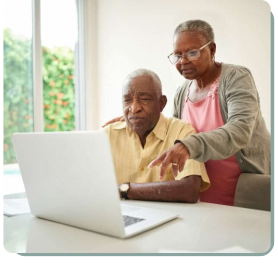 An elderly couple is sitting at a table using a laptop. The man, dressed in a yellow shirt, is seated directly in front of the laptop, focusing on the screen. The woman, wearing a pink top with a grey cardigan, stands beside him, pointing at the screen, seemingly offering assistance. They are in a bright room with a large window, through which green foliage is visible. The atmosphere is calm and supportive.