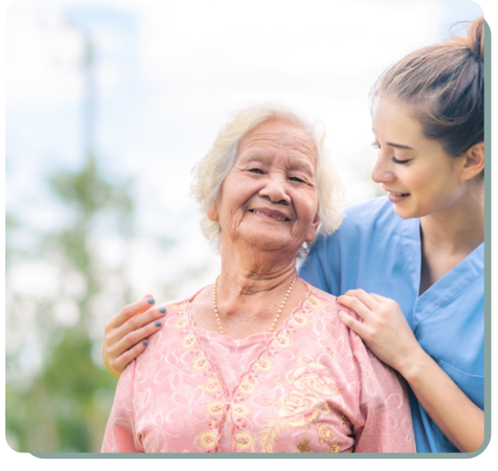 An elderly woman wearing a pink, embroidered top is standing outdoors, smiling warmly. She has white hair and is accessorized with a pearl necklace. A younger woman, dressed in a blue medical scrub top, stands beside her, gently placing her hands on the elderly woman's shoulders. The younger woman is smiling and looking affectionately at the elderly woman. The background is softly blurred with greenery, indicating they are in a garden or park on a bright, sunny day.