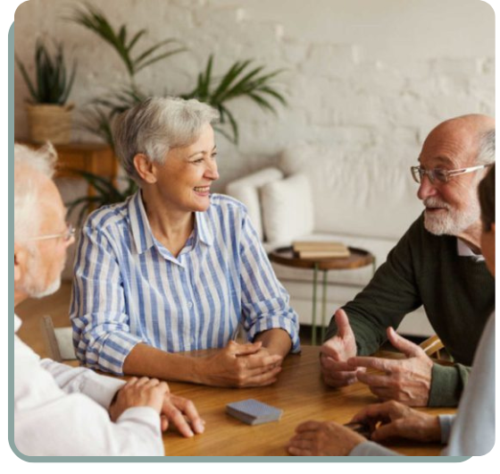 A group of four elderly people are seated around a wooden table in a cozy, well-lit room. The focus is on a woman with short gray hair, wearing a blue and white striped shirt, who is smiling and engaging in conversation with an older man with glasses and a beard, dressed in a dark green sweater. Two other men, partially visible, are also seated at the table, participating in the discussion. A deck of cards is placed on the table, suggesting they might be playing a game. The background features a neutral-toned wall and some potted plants, creating a warm and inviting atmosphere.