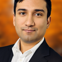 A man in a black suit and white collared shirt has a slight smile on his face and is standing in front of an orange background.