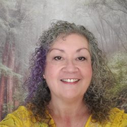 A therapist at Total Life, a smiling woman with curly gray hair, wearing a yellow top. She is set against a serene forest backdrop with trees and soft natural lighting.