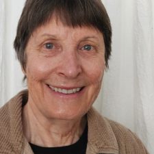 A portrait of an older woman, a therapist at Total Life, with short, dark hair, smiling warmly. She is wearing a tan corduroy jacket over a black top, set against a light curtain backdrop.