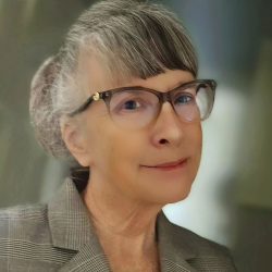 A professional portrait of an older woman - a therapist at Total Life - with gray hair styled in a bun, wearing glasses and a checkered blazer. She has a composed expression and is set against a softly blurred indoor background.