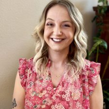 A therapist at Total Life with shoulder-length blonde hair styled in loose waves, wearing a pink floral blouse with ruffled sleeves. She is smiling warmly in an indoor setting with plants and a neutral background.