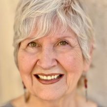 A smiling older woman - a therapist at Total Life - with short silver hair, wearing a gray top and dangling earrings. She is pictured against a softly blurred light-colored background.