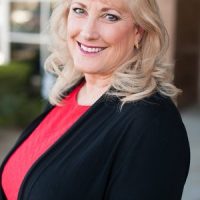 A therapist at Total Life with blonde hair styled in soft waves, wearing a black cardigan over a bright coral top. She is smiling warmly in an outdoor setting with a blurred building and greenery in the background.