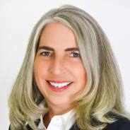 A therapist at Total Life with shoulder-length silver hair, wearing a white collared shirt and a black blazer. She is smiling brightly against a plain white background.