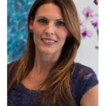 A portrait of a therapist at Total Life, a woman with long, light brown hair, wearing a navy blue lace top. She is smiling warmly in an indoor setting with floral and abstract decor in the background.