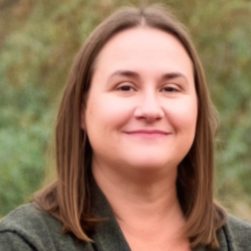 A portrait of a therapist at Total Life, a woman with shoulder-length brown hair, wearing a dark green top. She is smiling gently, set against a blurred natural outdoor background.