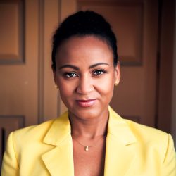 A professional headshot of a woman, a therapist at Total Life, with dark hair tied back, wearing a bright yellow blazer and a delicate necklace with a pendant. She is smiling softly, set against a warmly lit indoor background.