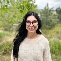 A therapist at Total Life, a smiling woman with long dark hair, wearing glasses and a beige sweater. She is outdoors in a lush, natural setting with trees and greenery in the background.