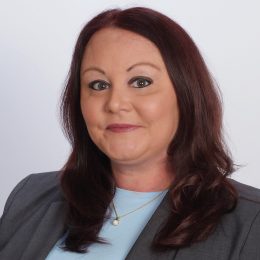 A professional headshot of a woman - a therapist at Total Life - with shoulder-length auburn hair, wearing a gray blazer, a light blue top, and a pearl necklace. She is smiling softly against a plain white background.