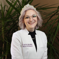 A therapist at Total Life with short, light lavender hair, wearing glasses and a white coat embroidered with her title. She is smiling warmly, seated in front of lush green plants in an indoor setting.