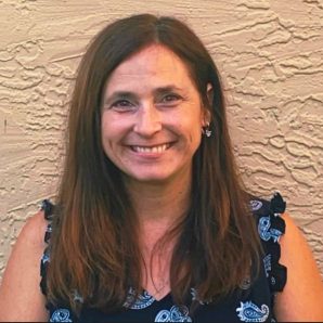 A therapist at Total Life, a smiling woman with long brown hair, wearing a dark patterned top with small floral designs. She is standing against a textured beige wall, creating a warm and natural backdrop.