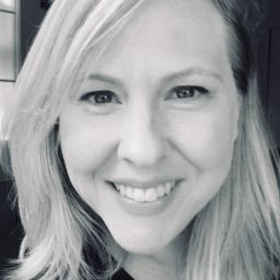 A black-and-white close-up portrait of a woman, a therapist at Total Life, with shoulder-length blonde hair, smiling warmly. The background features soft lighting from a window, giving a natural and inviting feel.