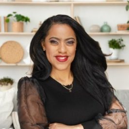 "A smiling woman with long dark hair, a therapist at Total Life, wearing a black top with sheer sleeves and a gold necklace. She is seated in a modern, well-lit indoor setting with shelves of plants and decor in the background.