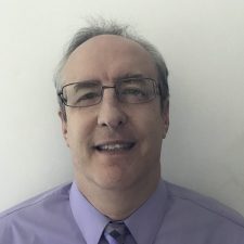 A professional headshot of a man - a therapist at Total Life - with short, slightly graying hair, wearing glasses, a lavender dress shirt, and a matching patterned tie. He is smiling softly against a plain white background.