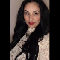 A therapist at Total Life with long, dark hair, wearing a cream lace top with a high neckline. She has a confident expression and is set against a neutral background.