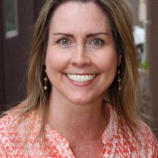 A professional headshot of a woman - a therapist at Total Life - with long blonde hair, wearing a gray blazer, a blue top, and a pearl necklace. She is smiling confidently in an indoor setting with a softly blurred background.