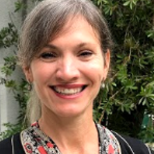 A smiling woman, a therapist at Total Life, with light gray hair tied back, wearing a patterned top with a black and red design. She is outdoors with greenery in the background, creating a natural and inviting setting.