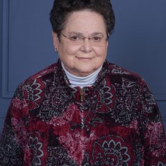 A therapist at Total Life with short, curly dark hair, wearing glasses, a patterned red and black zip-up jacket, and a white turtleneck. She is smiling softly against a plain blue background.