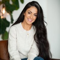 A therapist at Total Life with long, dark hair, wearing a light beige sweater and jeans. She is smiling warmly, seated in a chair with her hands clasped, in a modern indoor setting with plants in the background.