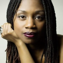 A close-up headshot of a woman - a therapist at Total Life - with long, styled locs, wearing deep burgundy lipstick and a confident expression. Her hand rests against her face, and she is adorned with a silver ring, set against a neutral background.