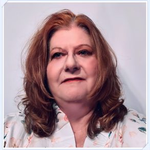 A portrait of a therapist at Total Life, a woman with shoulder-length auburn hair, wearing a floral-patterned blouse. She has a poised expression and is set against a plain white background with a subtle border framing the image.