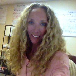 A photo of a woman, a therapist at Total Life, with long, curly blonde hair, smiling warmly. She is wearing a light pink shirt and is in an indoor setting with notes and charts visible on the walls in the background.