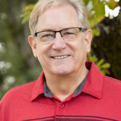 A therapist from Total Life, wearing glasses and a red polo shirt, smiling warmly in an outdoor setting with blurred greenery in the background.