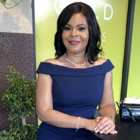 A smiling therapist from Total Life with shoulder-length dark hair, wearing an elegant navy off-shoulder dress and a pearl necklace, photographed indoors with a green and brown background.