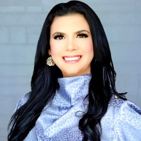 A smiling therapist from Total Life with long black hair, wearing intricate earrings and a lavender patterned blouse, photographed against a light blue background.