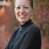 Portrait of a smiling therapist at Total Life with light skin and pulled-back hair, wearing a dark sweater in an outdoor autumn setting with a blurred background of warm orange and red foliage.