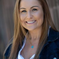 A smiling therapist from Total Life with long, straight light brown hair, wearing a white blouse, a denim jacket, and a turquoise pendant necklace, photographed outdoors with a blurred wooden background.