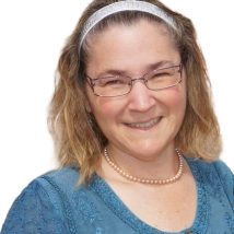 A smiling therapist from Total Life with shoulder-length blonde hair, wearing glasses, a pearl necklace, and a blue embroidered top, photographed against a white background.