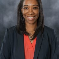 A therapist from Total Life dressed in a professional navy blazer and coral blouse, wearing a heart-shaped necklace and a friendly smile, set against a gray background.