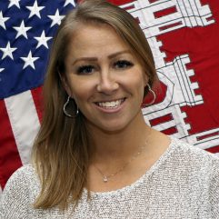 A therapist from Total Life with long blonde hair, smiling confidently. She is wearing a light sweater and a necklace, positioned in front of an American flag and another decorative flag, presenting a professional and patriotic demeanor.