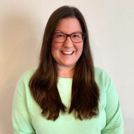 A smiling therapist from Total Life with long, straight brown hair, wearing glasses and a light green sweater, photographed against a neutral indoor background.