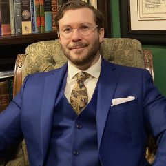 A therapist from Total Life dressed in a sharp blue suit with a paisley tie and pocket square, wearing glasses, seated in a vintage chair, and photographed indoors with a bookshelf and framed artwork in the background.