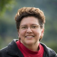 A therapist from Total Life. She is smiling and wearing glasses, a red turtleneck sweater, and a dark jacket. The outdoor background showcases soft, blurred greenery, adding a natural and warm tone to the photo.