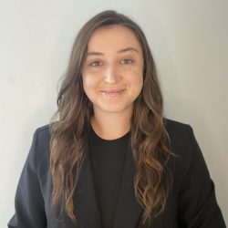 A smiling therapist from Total Life with long wavy brown hair, wearing a black blazer and top, photographed indoors against a light gray background.