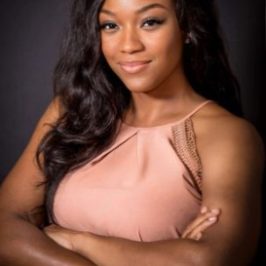 A confident therapist from Total Life with long, wavy dark hair, wearing a light pink sleeveless top, photographed against a dark background with arms crossed.