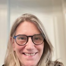 A smiling therapist from Total Life with light blonde hair, wearing glasses, photographed indoors with a neutral background.