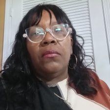 A therapist from Total Life with wavy black hair, wearing decorative glasses, hoop earrings, and layered clothing, photographed indoors against a light-colored door background.