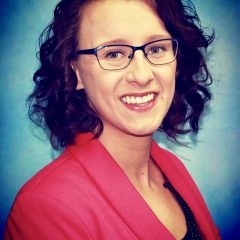 A therapist from Total Life. She is wearing glasses and a bright pink blazer over a dark top, smiling warmly. Her curly hair frames her face, and the background is a gradient of blue tones.