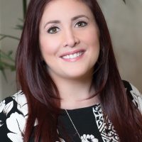 A smiling therapist from Total Life with long, straight auburn hair, wearing a black and white floral-patterned top, photographed indoors with greenery in the background.